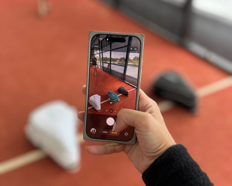 Person holding a smartphone taking a photo of three padel backapcks  on an orange padel court.
